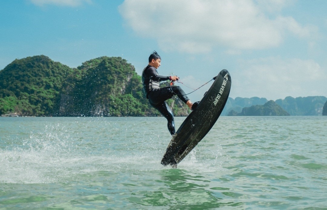 Người đàn ông trượt nước, mặc đồ bảo hộ, cưỡi ván trượt buồm, hành động ấn tượng trên vịnh nước xanh, núi non xa xa.