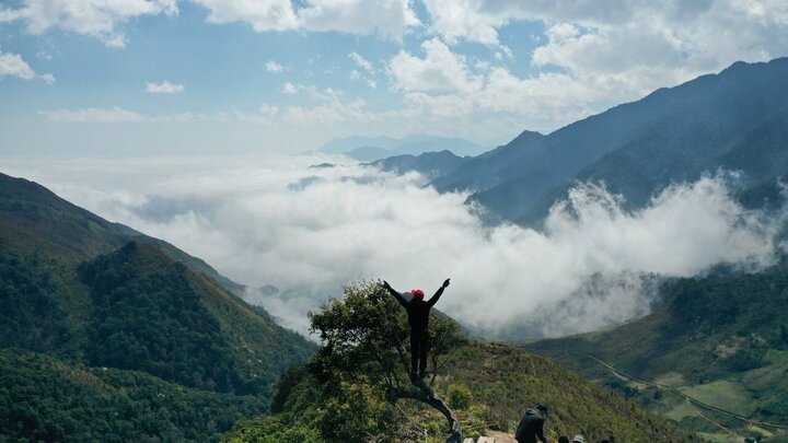 Người leo núi giơ tay chiến thắng trên đỉnh đồi, nhìn ra khung cảnh thiên nhiên hùng vĩ với mây mù và dãy núi trùng điệp.