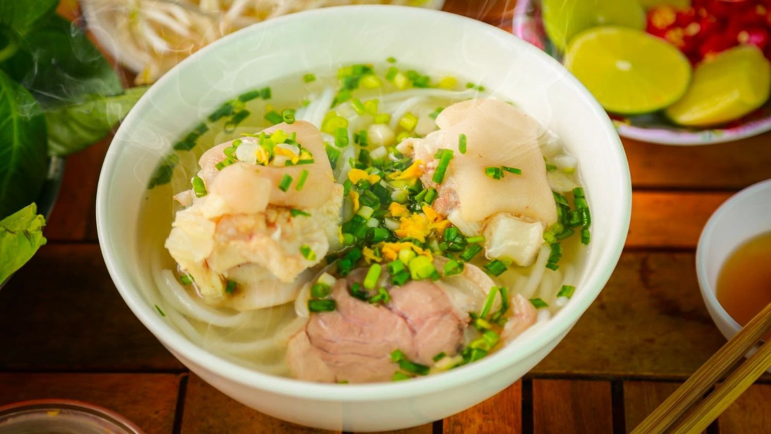 Bát phở nóng hổi với những miếng thịt bò thái lát, hành lá, và rau thơm, bốc khói thơm phức trên bàn gỗ.