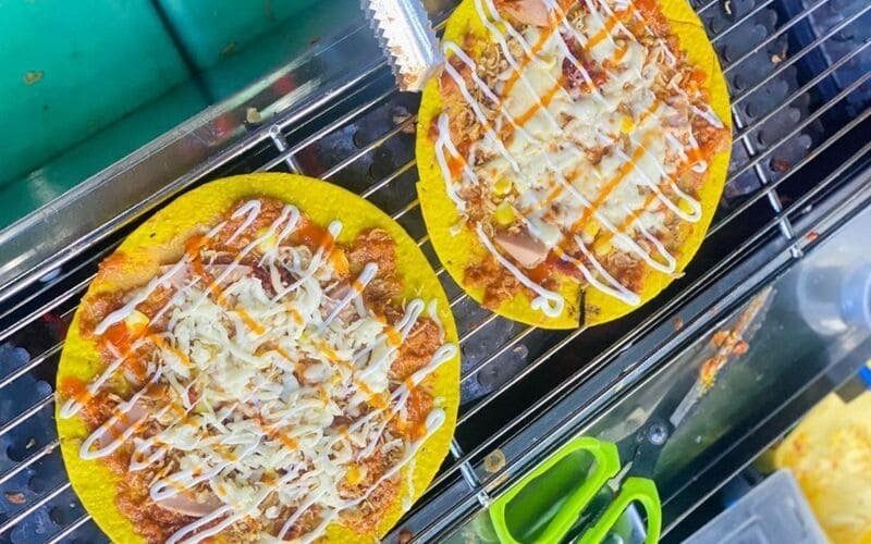 Hai đĩa bánh tráng nướng đầy màu sắc, phủ đầy phô mai và các loại sốt, đang nướng trên vỉ, bày trên bàn.