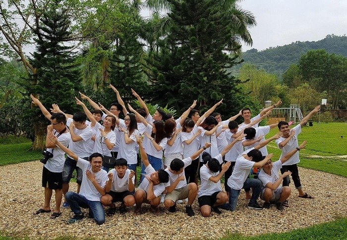 Khám Phá Đắk Lắk: Hành Trình Team Building Đầy Thú Vị Và Khác Biệt 2 Nhóm lớn các bạn trẻ mặc áo thun trắng tạo dáng vui nhộn giữa thiên nhiên xanh tươi và đồi núi xanh mát phía sau.