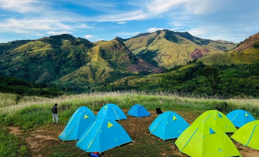 Những chiếc lều màu xanh dựng trên đồng cỏ xanh tươi, phía trước là dãy núi xanh mượt vào một ngày trời trong xanh.