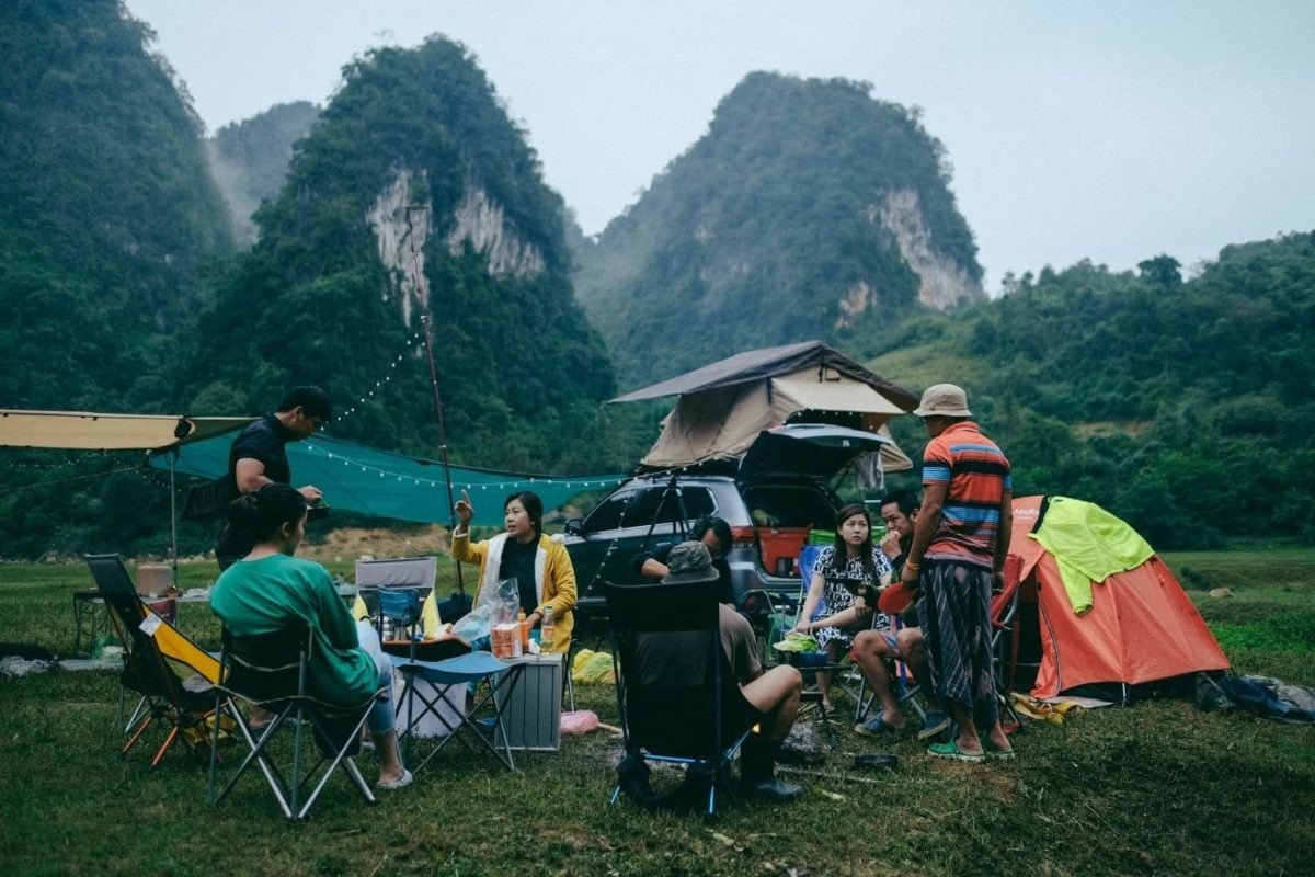 Một nhóm bạn đang cắm trại ngoài trời với xe hơi, lều trại và núi non hùng vĩ ở phía sau tạo nên khung cảnh tuyệt đẹp.