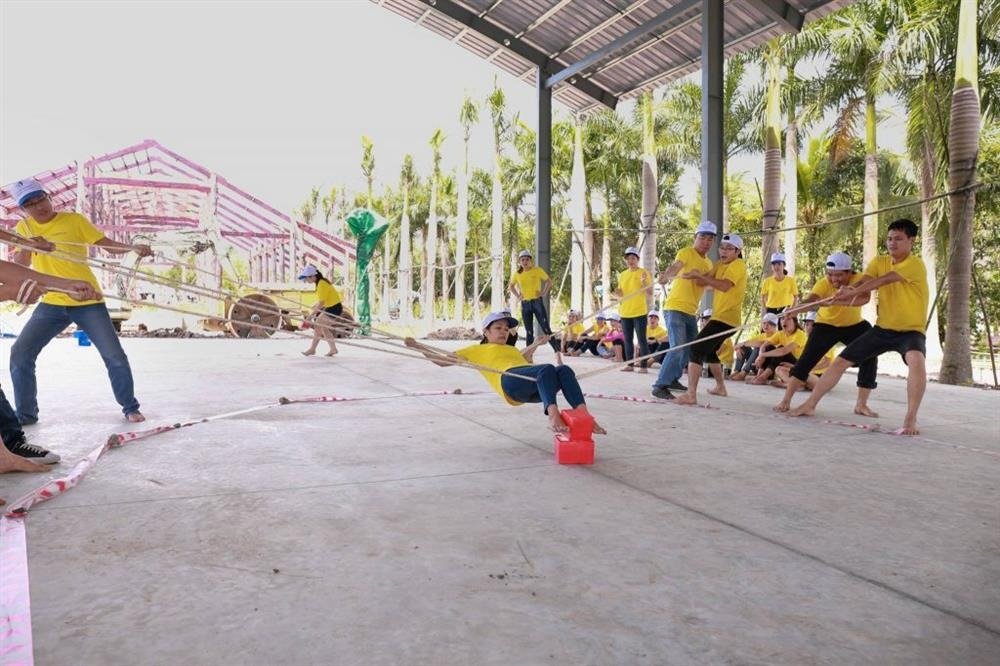 Các thành viên trong đội đeo áo thun màu vàng đang tham gia trò chơi kéo co tập thể ngoài trời tại một khu sân có mái che.
