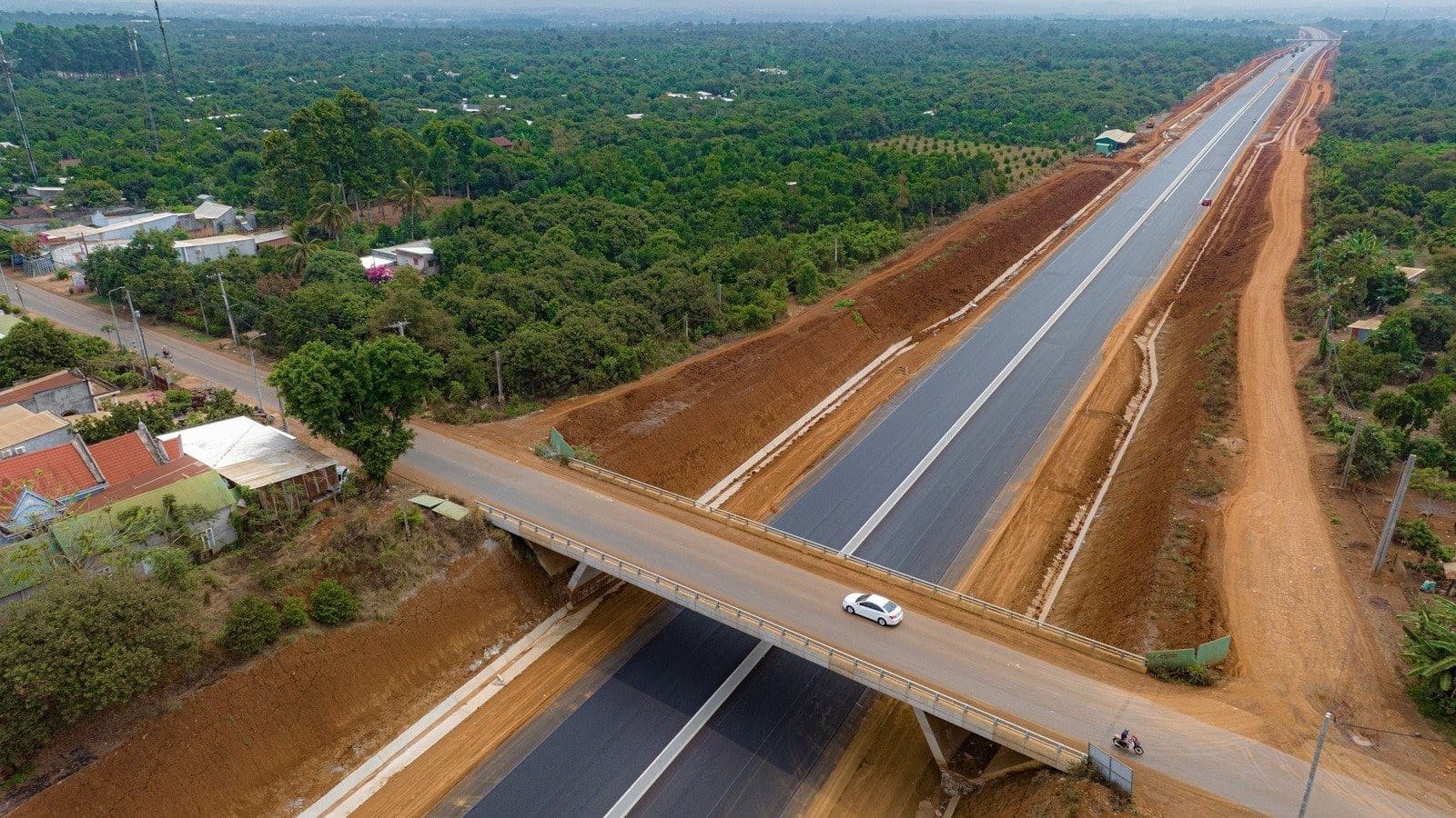 Hình ảnh một cây cầu bắc qua con đường cao tốc mới đang được xây dựng, với khung cảnh xanh tươi của vùng nông thôn xung quanh.