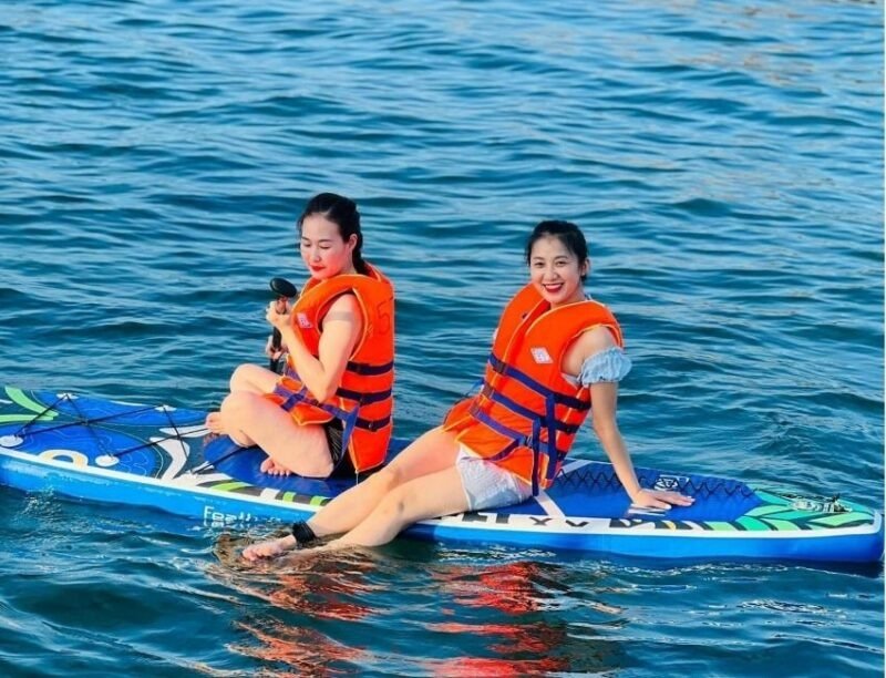 Hai cô gái mặc áo phao cam ngồi trên ván chèo trên mặt nước xanh, tận hưởng thời gian chèo ván và thư giãn ngoài trời.