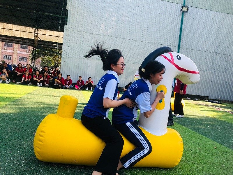 Hai người phụ nữ tham gia cuộc thi trên sân cỏ, cưỡi một phao bơm hơi hình con vật, tạo không khí vui tươi, sôi động.