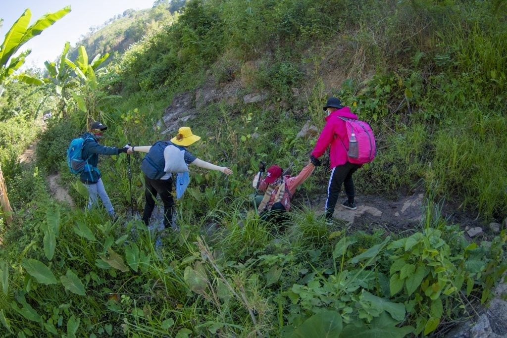 Một nhóm người leo núi giúp đỡ nhau qua địa hình dốc tại khu vực rừng nhiệt đới xanh tươi.