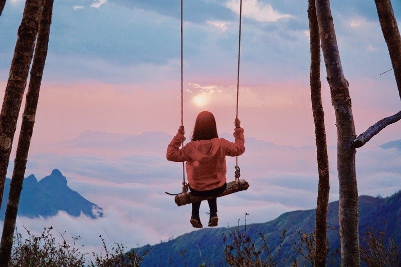 Người ngồi trên xích đu giữa khung cảnh hoàng hôn núi non thơ mộng, thiên nhiên đẹp và huyền bí, nền trời mây bồng bềnh.