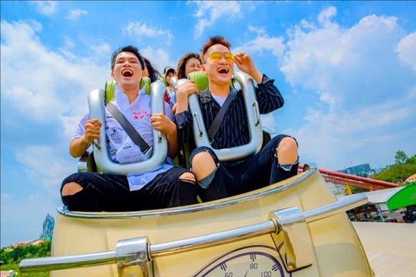 Hai người đàn ông đang cười lớn khi ngồi trên tàu lượn siêu tốc dưới bầu trời xanh với mây trắng.