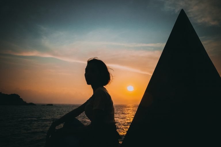 Người phụ nữ ngắm hoàng hôn trên biển, khung cảnh yên bình với bóng dáng tạo thành từ ánh sáng hoàng hôn.