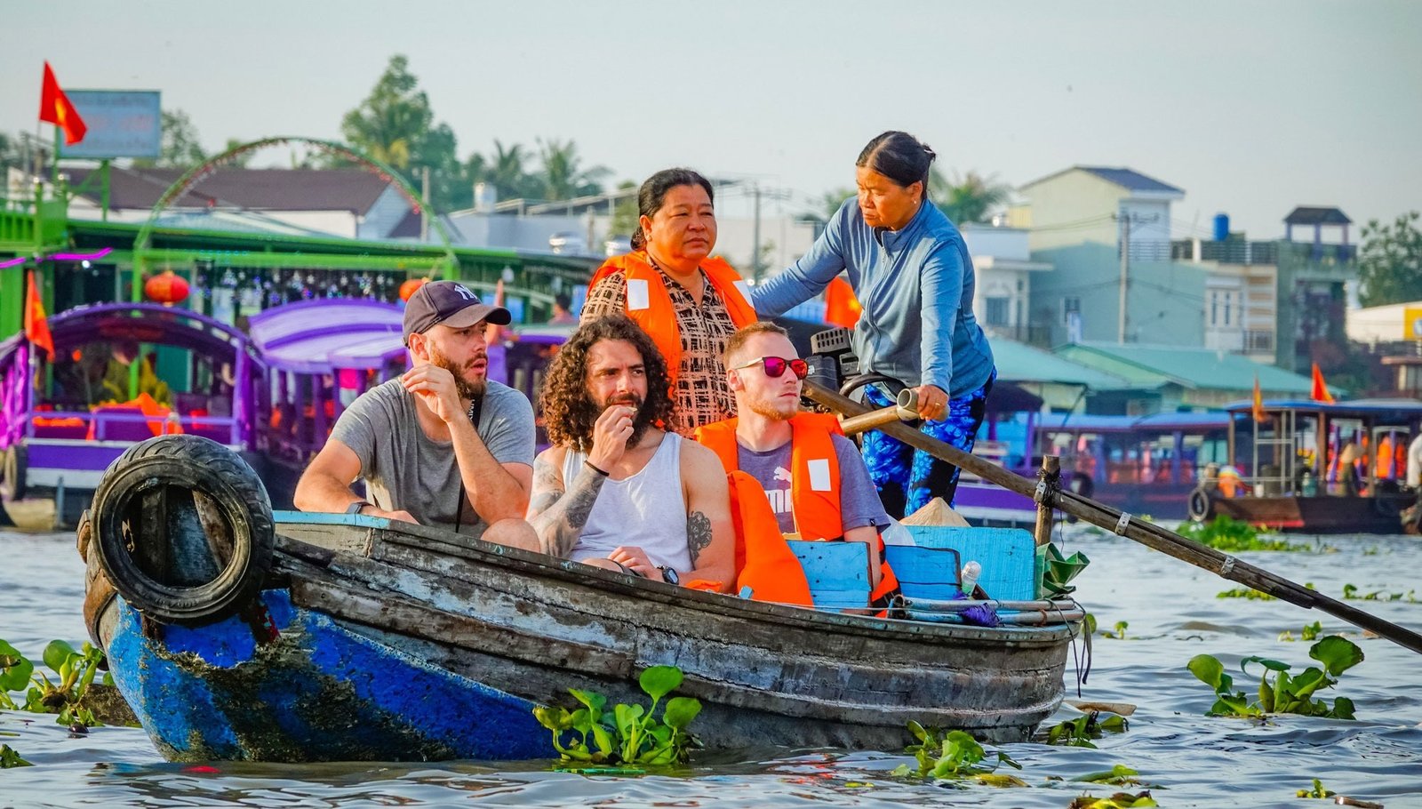 Du Lịch Quốc Tế Là Gì? Khám Phá Các Loại Hình Du Lịch Quốc Tế Phổ Biến Hiện Nay 3 Du khách ngồi trên thuyền chèo truyền thống, tham quan chợ nổi ở vùng sông nước Việt Nam với áo phao an toàn.