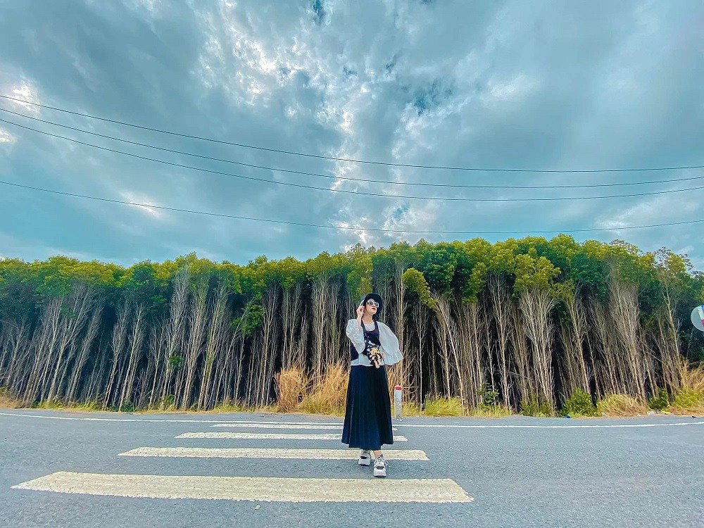 Cô gái đứng trên vạch kẻ đường giữa trời xanh và khu rừng sậy cao, tạo dáng chụp hình dưới bầu trời mây.