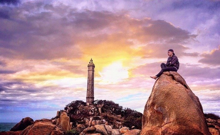 Người đàn ông ngồi trên tảng đá lớn ngắm bình minh bên ngọn hải đăng cổ giữa khung cảnh thiên nhiên hùng vĩ.