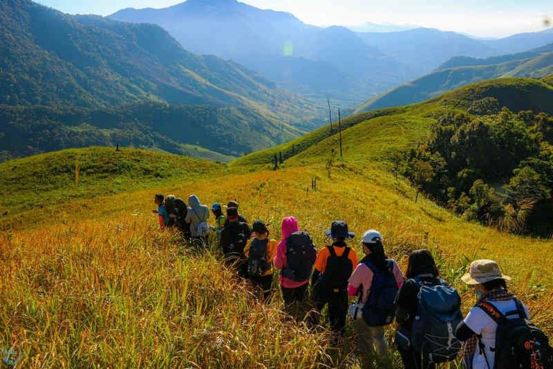 Nhóm du khách trekking xuống đồi cỏ xanh giữa khung cảnh núi non hùng vĩ và thung lũng dưới bầu trời xanh.