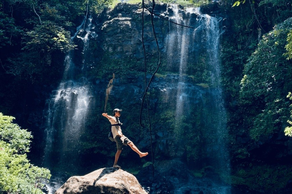 Người đàn ông đang đu dây trước thác nước hùng vĩ trong rừng nhiệt đới xanh mướt, tạo cảm giác phiêu lưu mạo hiểm.