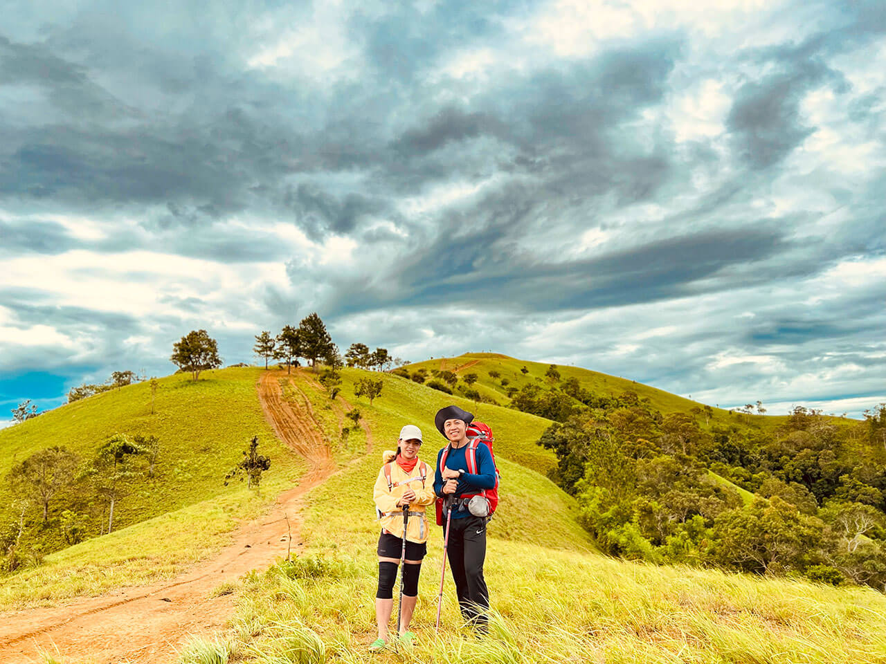 Hai người leo núi đứng trên đồi cỏ xanh mướt dưới bầu trời u ám, sẵn sàng cho chuyến hành trình phiêu lưu.