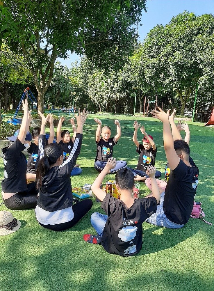 Team Building Học Sinh: Chìa Khóa Phát Triển Kỹ Năng Mềm Quan Trọng 3 Nhóm trẻ em ngồi thành vòng tròn trên bãi cỏ xanh, giơ tay lên cao dưới bóng cây trong công viên đầy nắng.