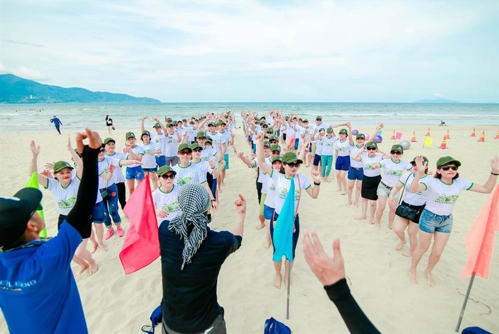 Nhóm người tham gia trò chơi team building sôi động trên bãi biển, mang áo thun trắng và đội mũ xanh, cùng nhau giơ tay chào.