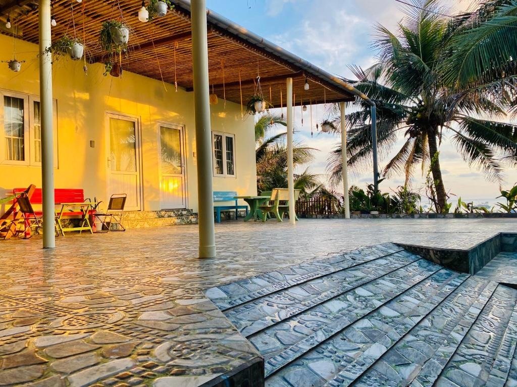 Nhà nghỉ nhỏ màu vàng với sân hiên nhìn ra biển, có bàn ghế đầy màu sắc, cây cọ và ánh nắng hoàng hôn.