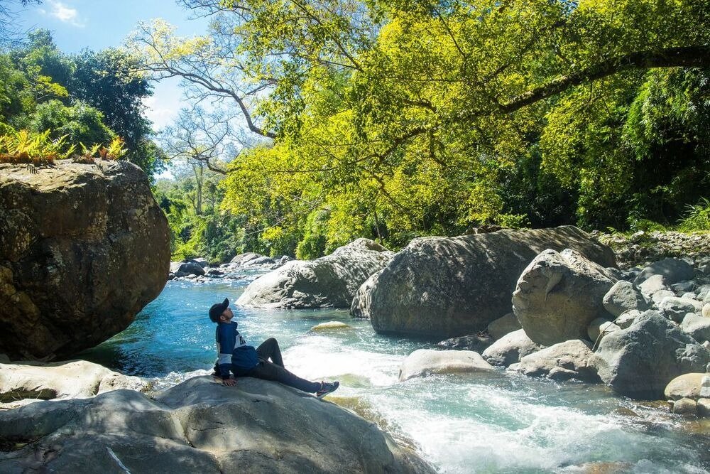 Người ngồi thư giãn bên bờ suối, cây cối tươi tốt và đá lớn, tạo nên khung cảnh thiên nhiên yên bình.