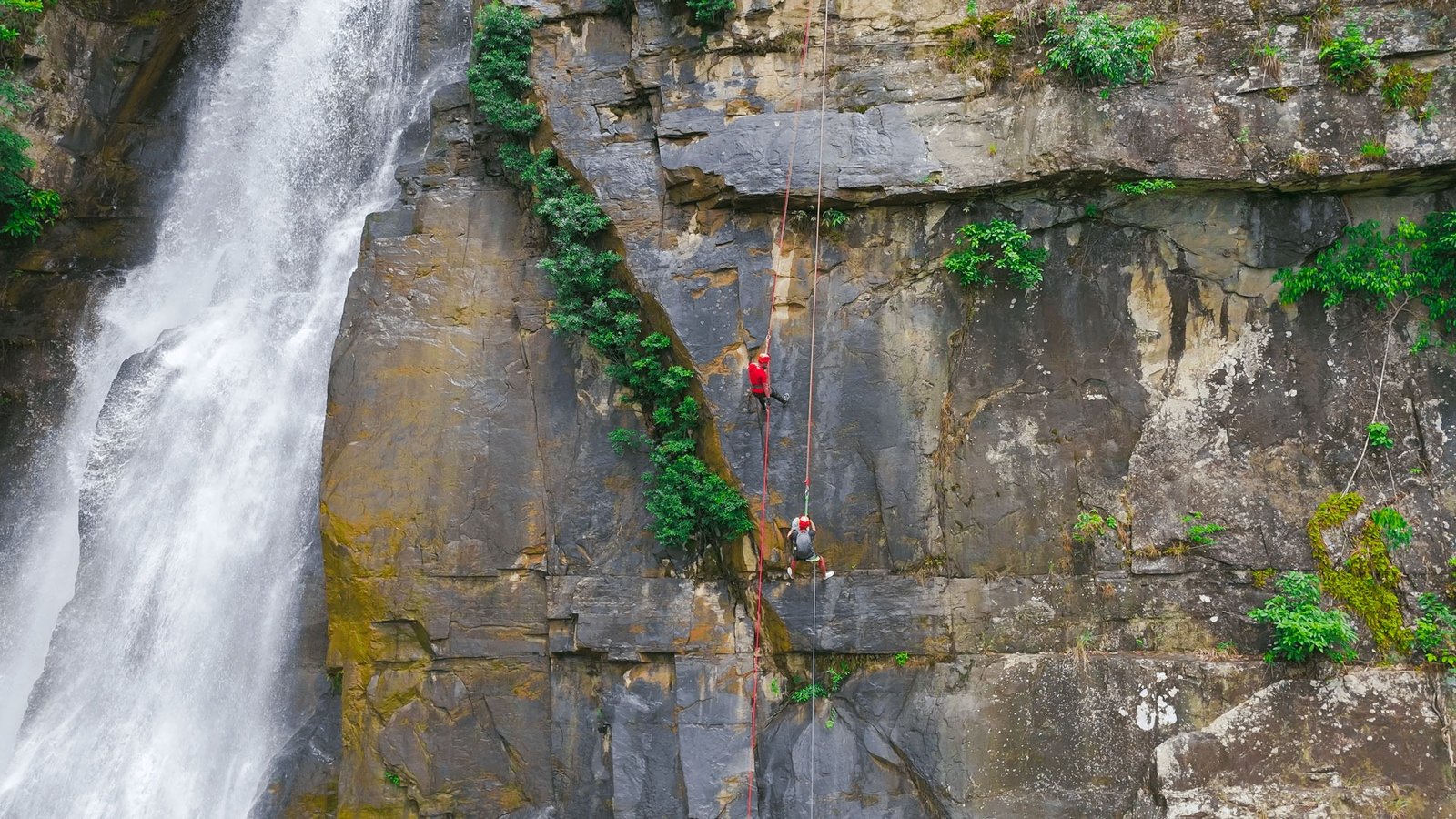 Hai người leo núi đang trèo lên bên cạnh một thác nước hùng vĩ, với dây thừng và thiết bị an toàn.