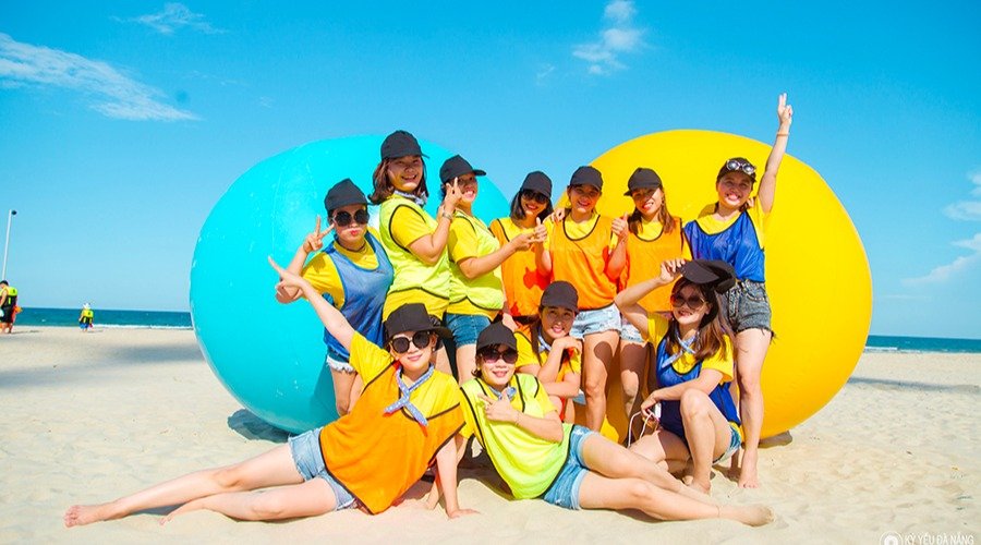Nhóm các cô gái đang mặc áo phông màu sắc, đội mũ và chụp ảnh bên các quả bóng lớn trên bãi biển nắng vàng.