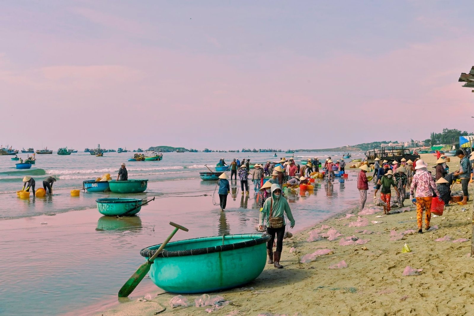Ngư dân đang làm việc trên bãi biển với những chiếc thuyền thúng và thuyền đánh cá ở xa, dưới bầu trời bình minh.