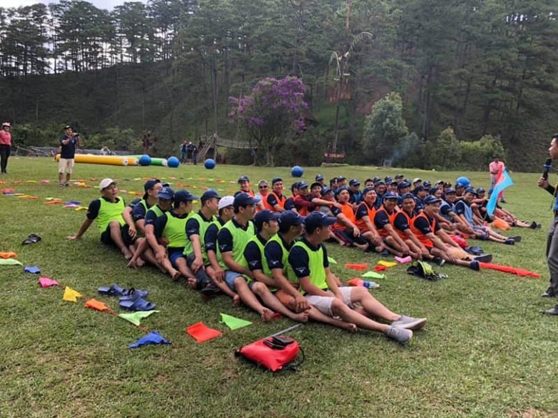 Team Building Amazing Race Đà Lạt: Trải Nghiệm Độc Đáo Cho Doanh Nghiệp 3 Nhóm người mặc áo đồng phục màu sắc đang tham gia hoạt động ngoài trời trên bãi cỏ xanh với cây cối xung quanh.