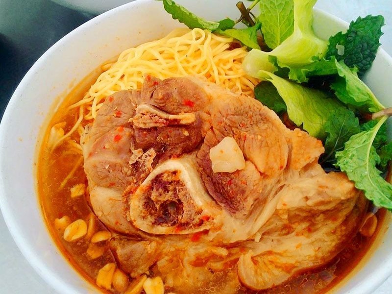 Bát bún bò Huế đầy hấp dẫn, với miếng giò heo lớn, nước lèo đậm đà và rau sống xanh mướt bên cạnh.