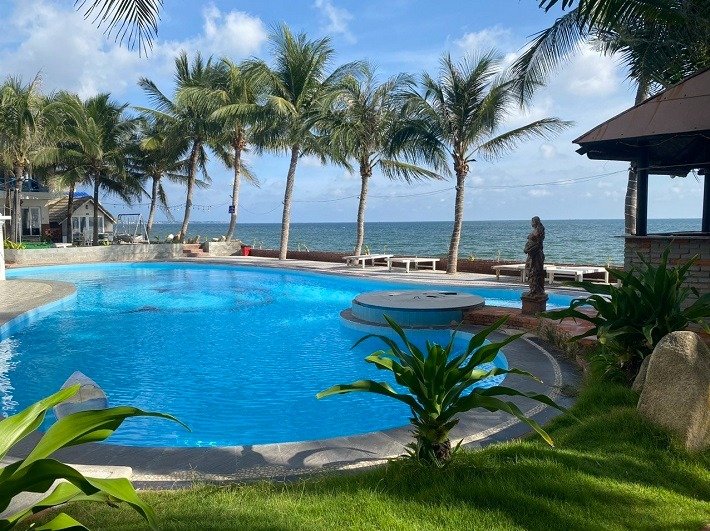 Hồ bơi ngoài trời xanh biếc với hàng dừa, ghế tắm nắng, bức tượng và biển xanh xa xỉ dưới bầu trời xanh.