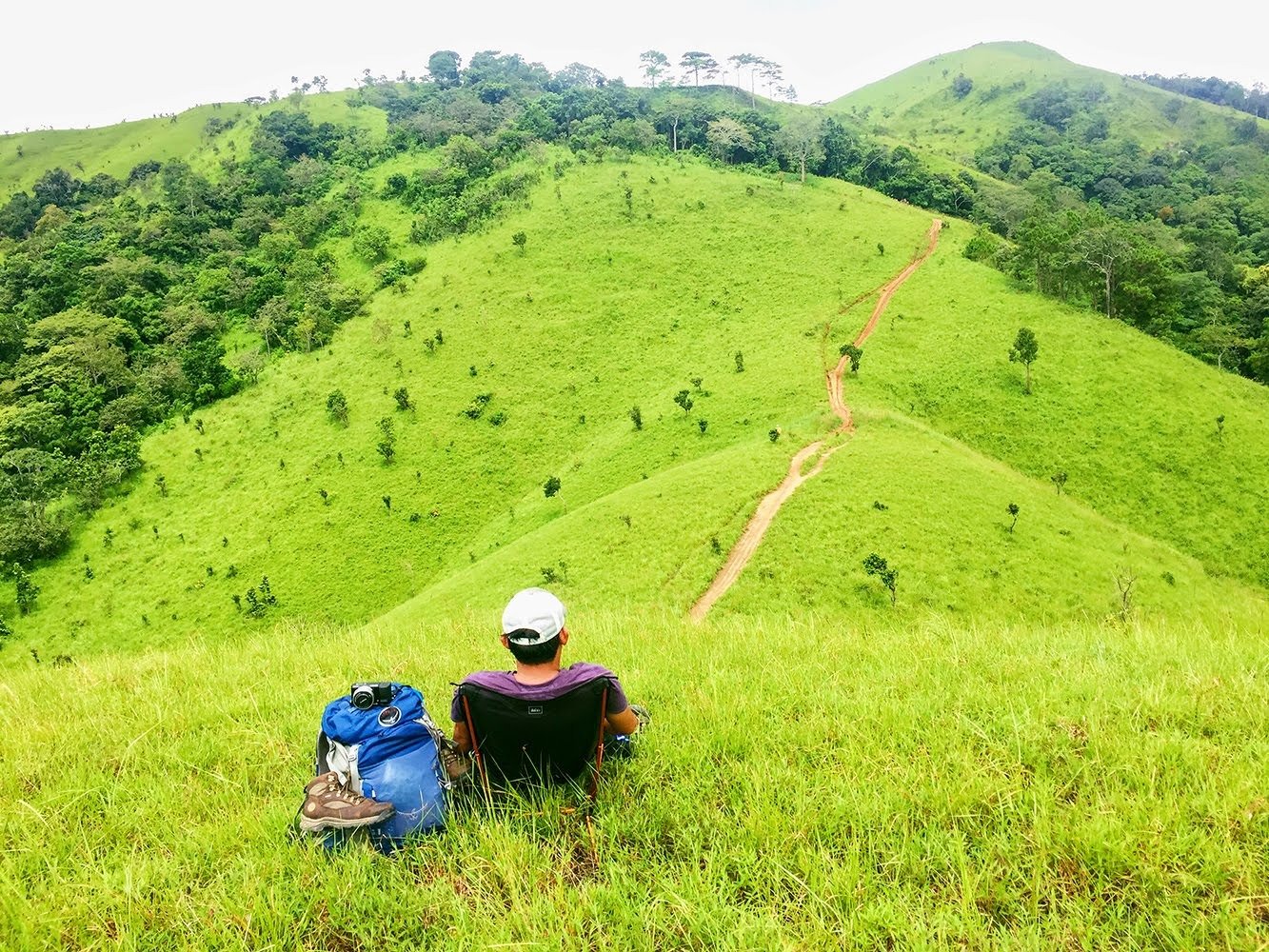 Người đi bộ ngồi nghỉ trên đồng cỏ xanh mướt, đối diện đoạn đường mòn đỏ dẫn qua những ngọn đồi dốc đầy cây cối.