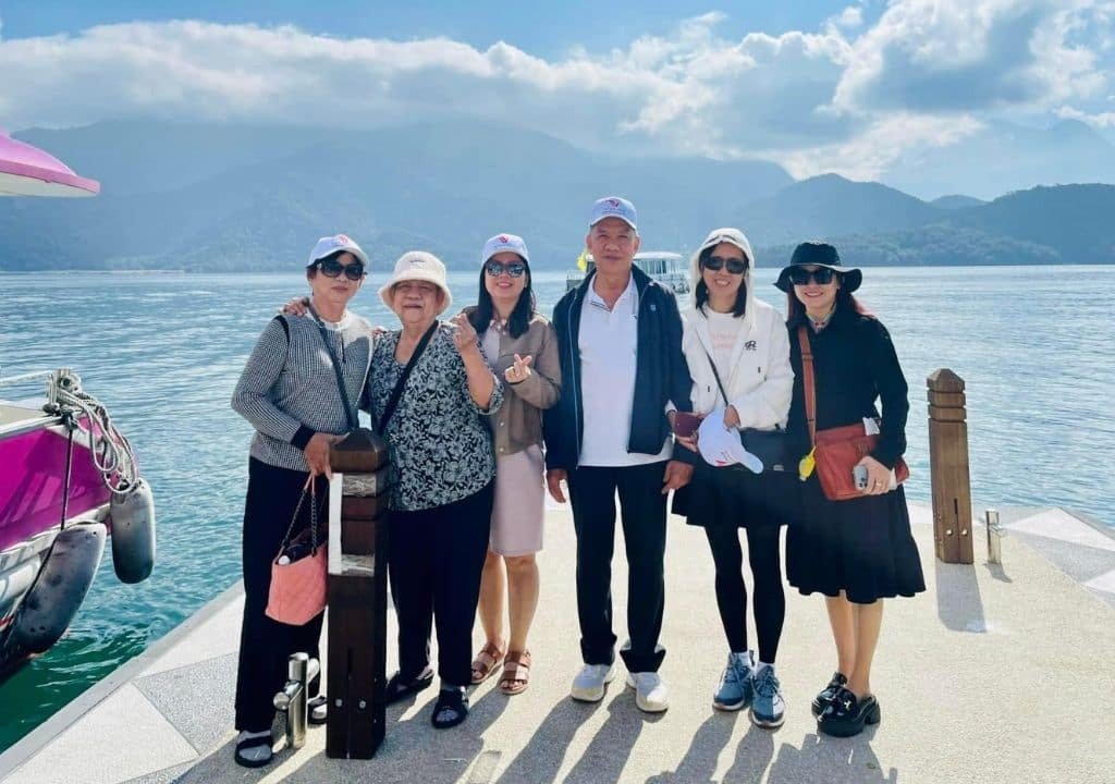 Nhóm người đứng bên hồ trong một chuyến đi chơi, với núi đồi và mây trắng làm nền phong cảnh hữu tình.