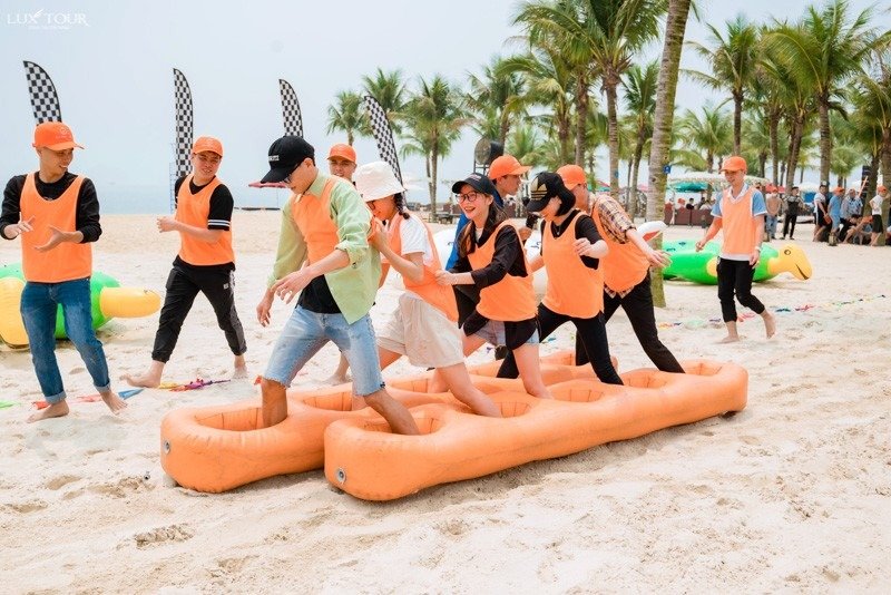 Team Building Vũng Tàu: Trải Nghiệm Thú Vị Tại Thành Phố Biển Tuyệt Đẹp 3 Nhóm người tham gia hoạt động team building vui nhộn trên bãi biển, mặc áo cam và đội mũ, vui chơi trên cát.