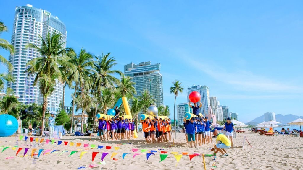 Nhóm người mặc áo màu sắc tham gia trò chơi ngoài trời trên bãi biển với nền tòa nhà hiện đại.