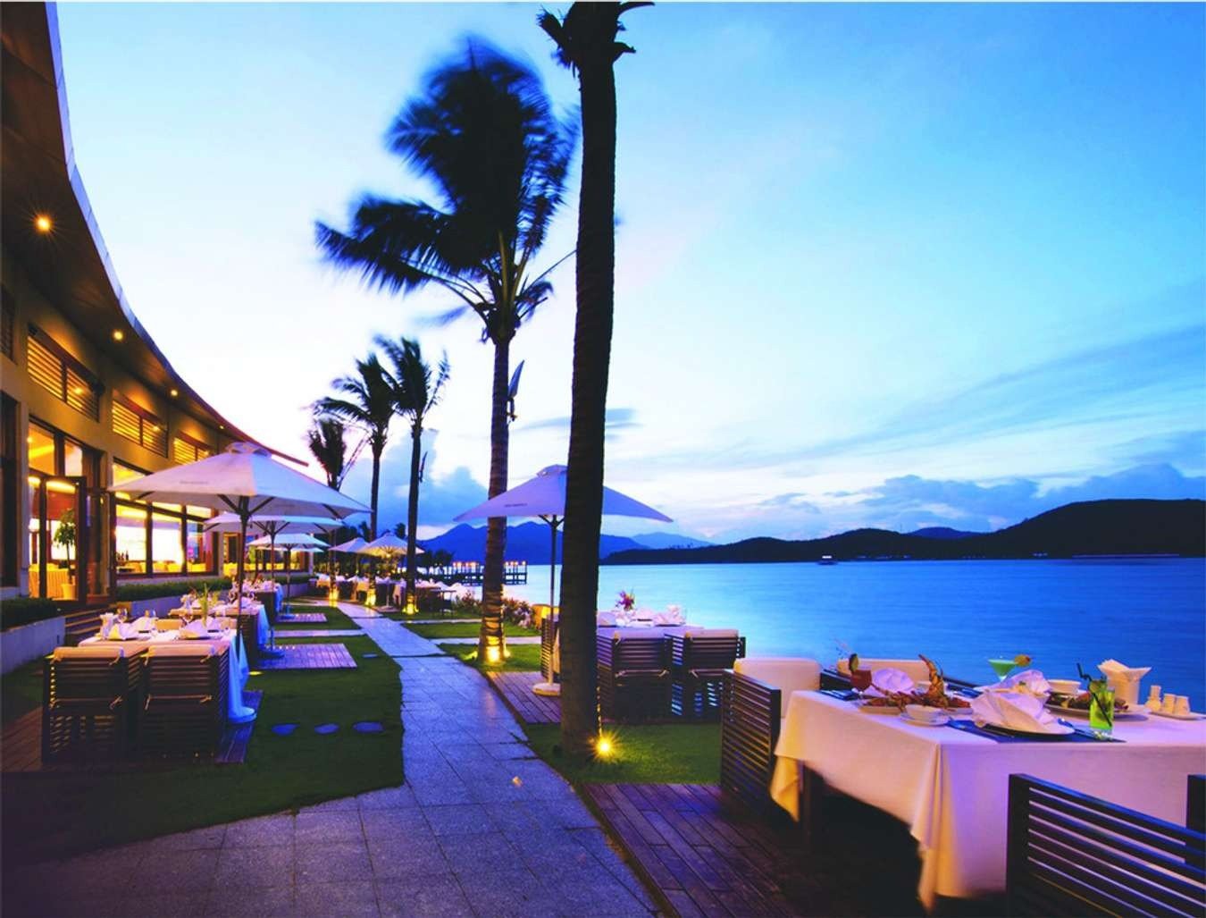 Khu vực ăn uống ngoài trời sang trọng cạnh biển với cảnh hoàng hôn tuyệt đẹp, cây cọ và bàn ghế được bài trí tinh tế.