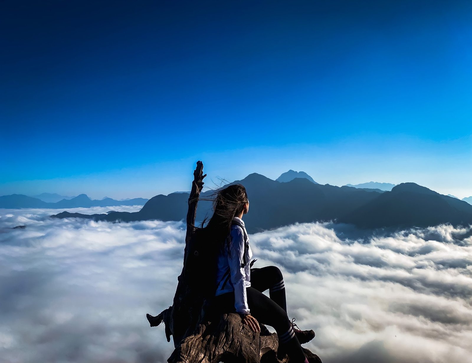 Người phụ nữ ngồi trên đỉnh núi ngắm nhìn biển mây bao quanh dãy núi hùng vĩ dưới bầu trời xanh biếc.