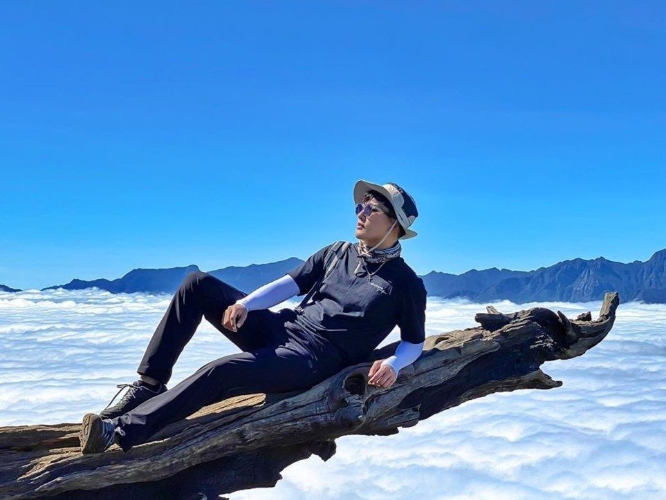 Người đàn ông nằm thư giãn trên cành cây trên cao, có nền là biển mây trắng và núi non hùng vĩ trong bầu trời xanh.