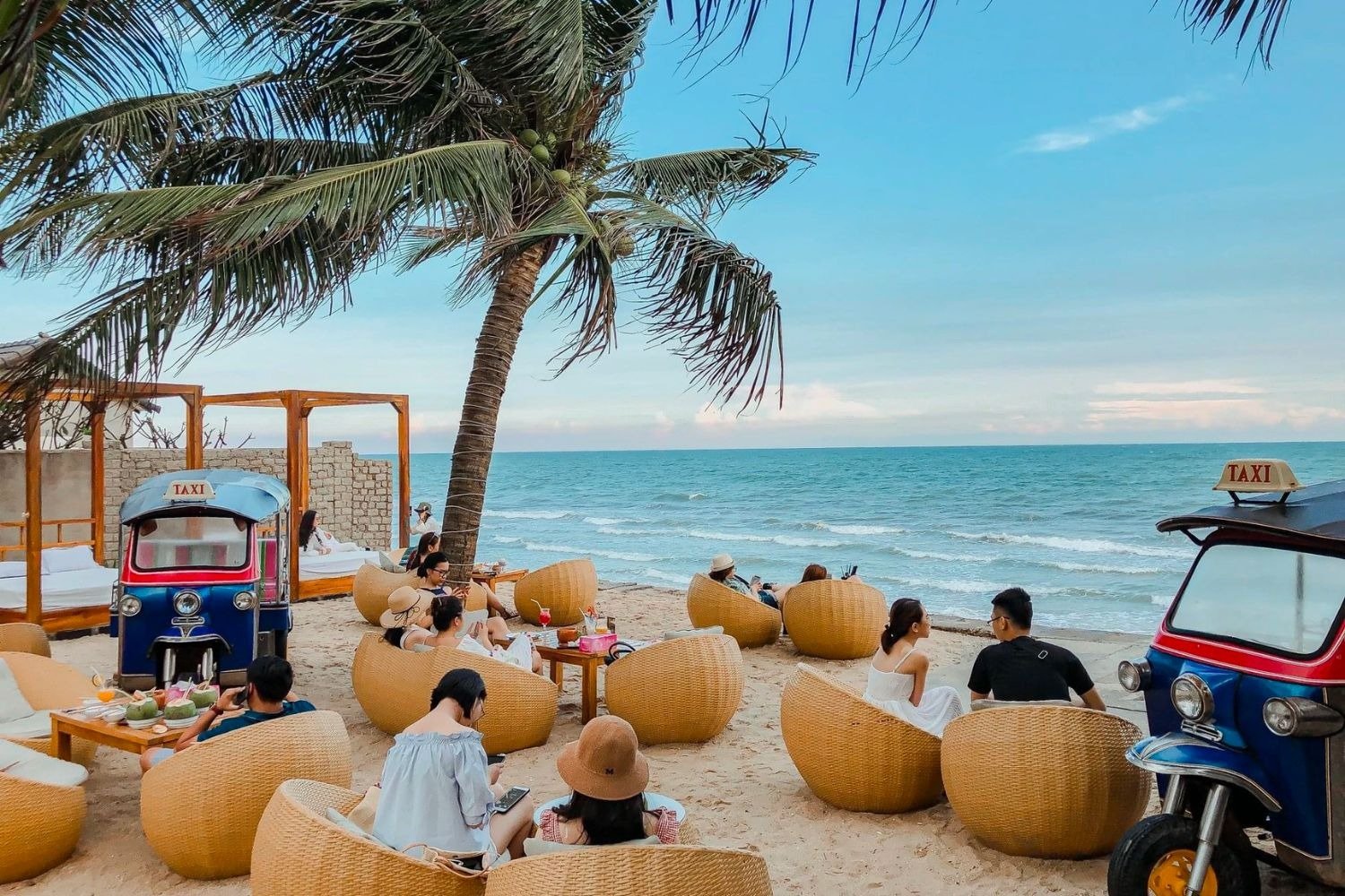Du khách tận hưởng hoàng hôn bên bờ biển xanh với đồ uống mát lạnh, ghế mây êm ái và không gian yên bình, thơ mộng.