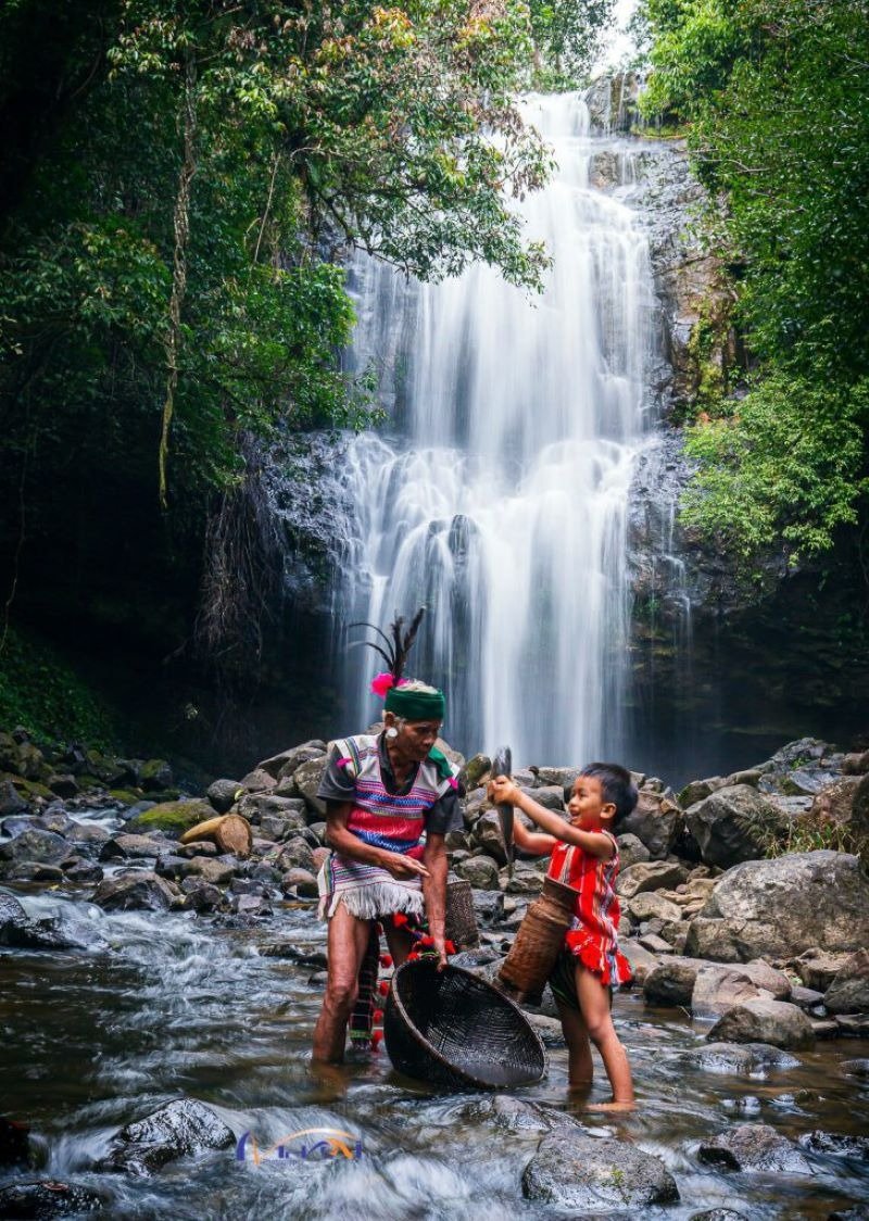 Người lớn và trẻ em với trang phục truyền thống bắt cá bên thác nước, xung quanh là cây cối xanh tươi.