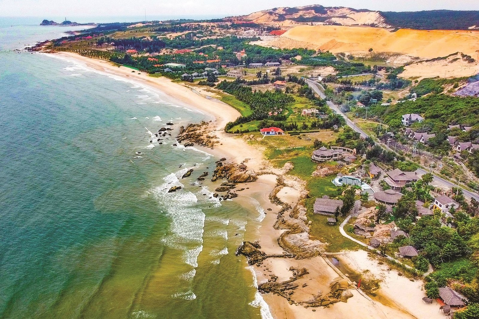 Cảnh quan bãi biển dài với các khu nhà nghỉ và resort bên bờ biển, phía sau là các đồi cát và rừng cây xanh dày đặc.