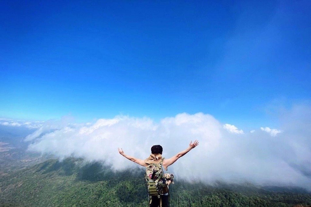 Chinh Phục Tây Nguyên: Top 5 Tour Trekking Đỉnh Cao Không Thể Bỏ Lỡ 3 Một người leo núi đứng trên đỉnh, giang tay trước bầu trời xanh và đám mây trắng, cảm nhận vẻ đẹp thiên nhiên.