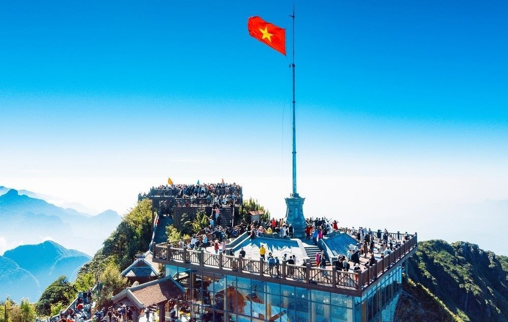 Mô Hình Du Lịch Giải Trí: Xu Hướng Hấp Dẫn Khách Du Lịch Hiện Đại 5 Đỉnh Fansipan với lá cờ Việt Nam tung bay, nơi có tầm nhìn toàn cảnh núi non hùng vĩ dưới bầu trời trong xanh.