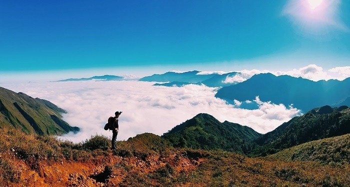 Người leo núi đứng trên sườn đồi, ngắm nhìn biển mây trải dài dưới núi đồi xanh biếc với trời xanh trong vắt.
