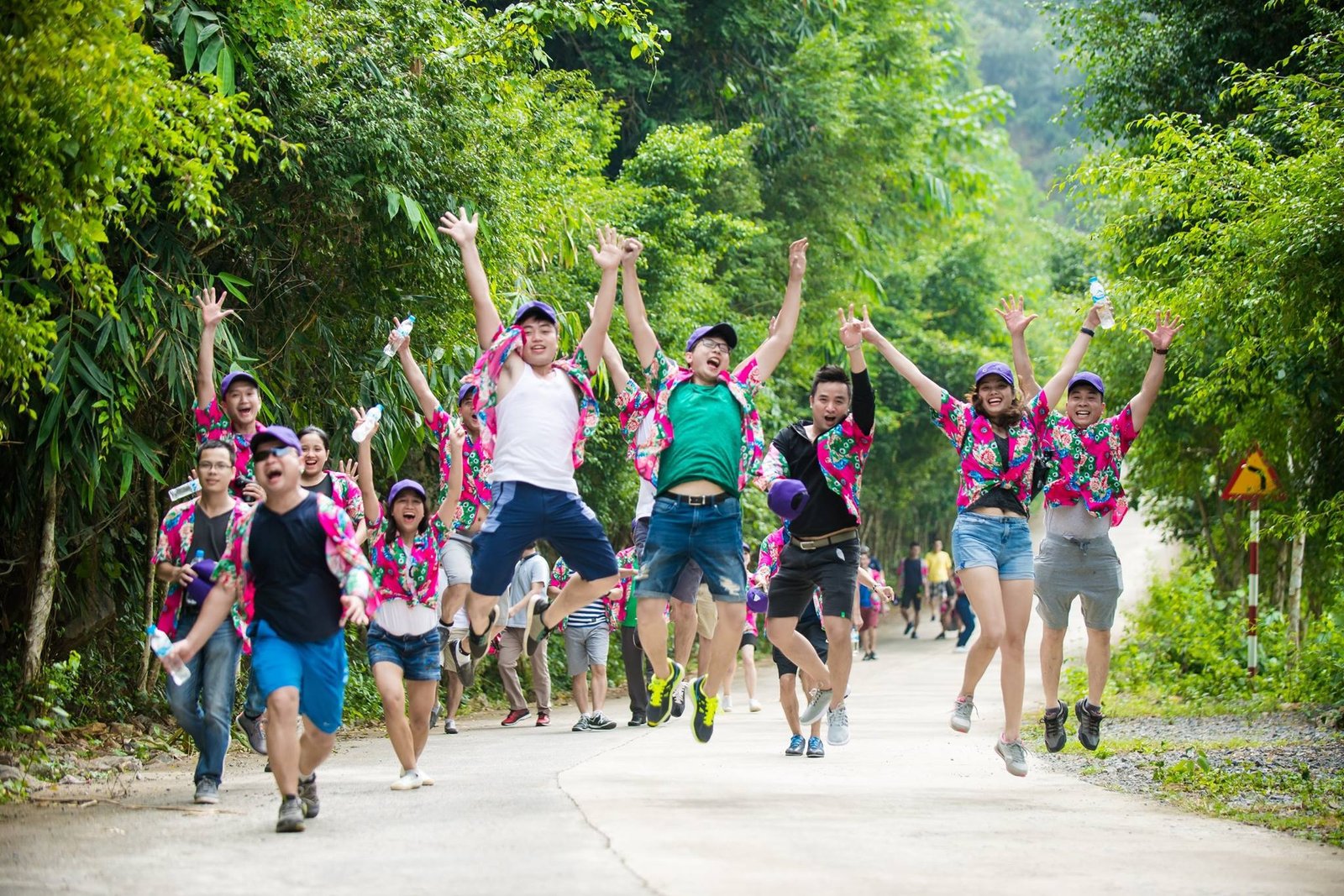 Một nhóm bạn trẻ mặc áo hoa rực rỡ, đội mũ tím, nhảy lên vui tươi giữa con đường rợp bóng cây xanh mát, thể hiện niềm vui và sự đoàn kết.