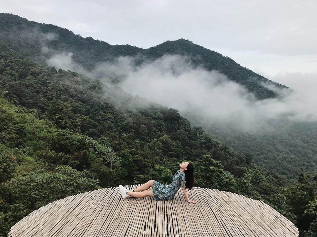 Người phụ nữ thư giãn trên nền tre với phong cảnh núi đồi phủ sương mù, tạo nên khung cảnh thiên nhiên ấn tượng.