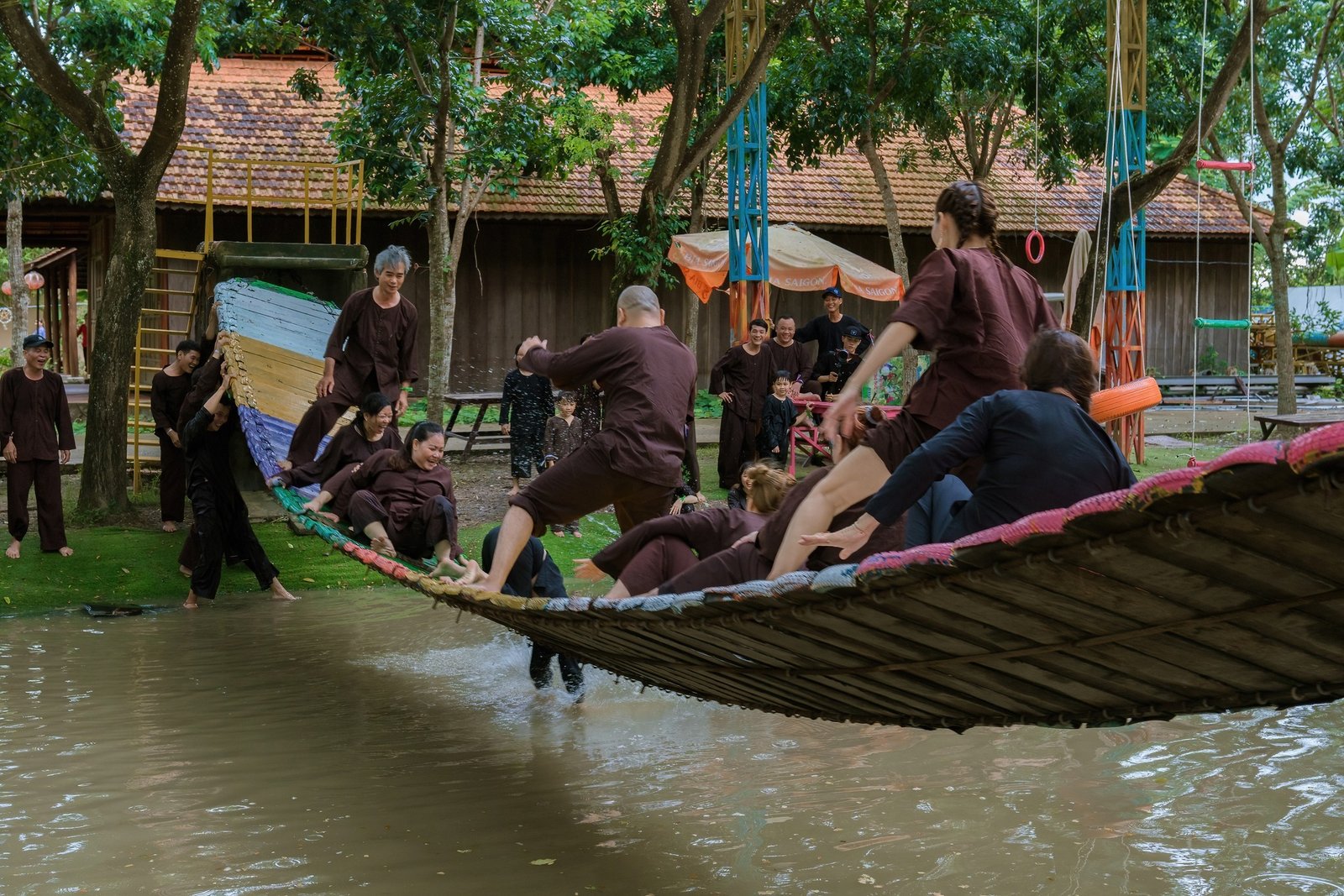 Một nhóm người mặc đồ truyền thống, chơi cùng nhau trên một cây cầu tre bắc qua hồ tại một khu vực nông thôn.