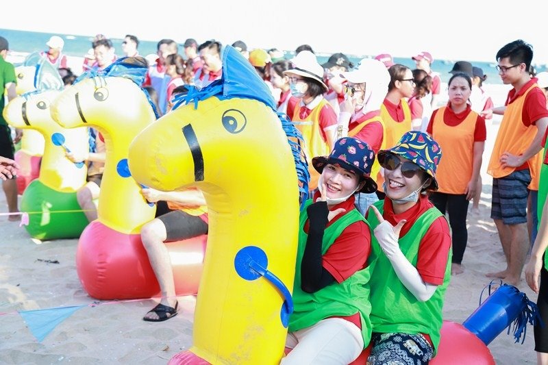 Hình ảnh các nhân viên công ty tham gia trò chơi tập thể trên bãi biển, ngồi trên ngựa hơi màu vàng sáng.