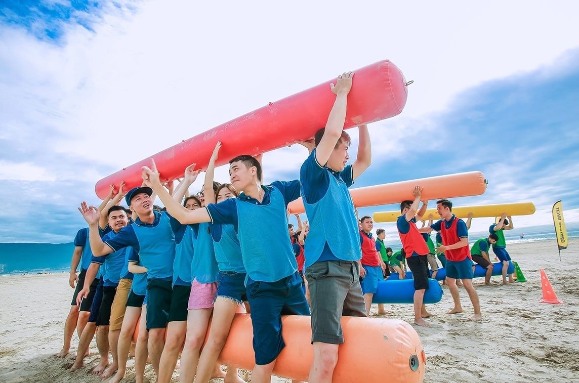 Nhóm người mặc áo xanh tham gia hoạt động teambuilding đầy nhiệt huyết trên bãi biển, mang ống lớn màu sắc tươi sáng.