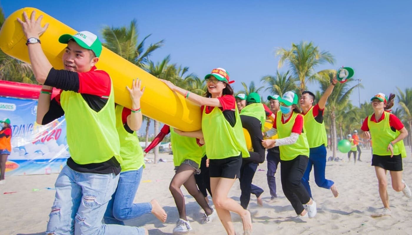 Nhóm người tham gia trò chơi teambuilding sôi động trên bãi biển, mặc áo phông đỏ và mặc áo vest xanh, đội mũ bảo hiểm.